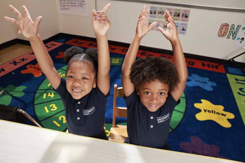 Two young students sitting at a desk raising their hands in the air